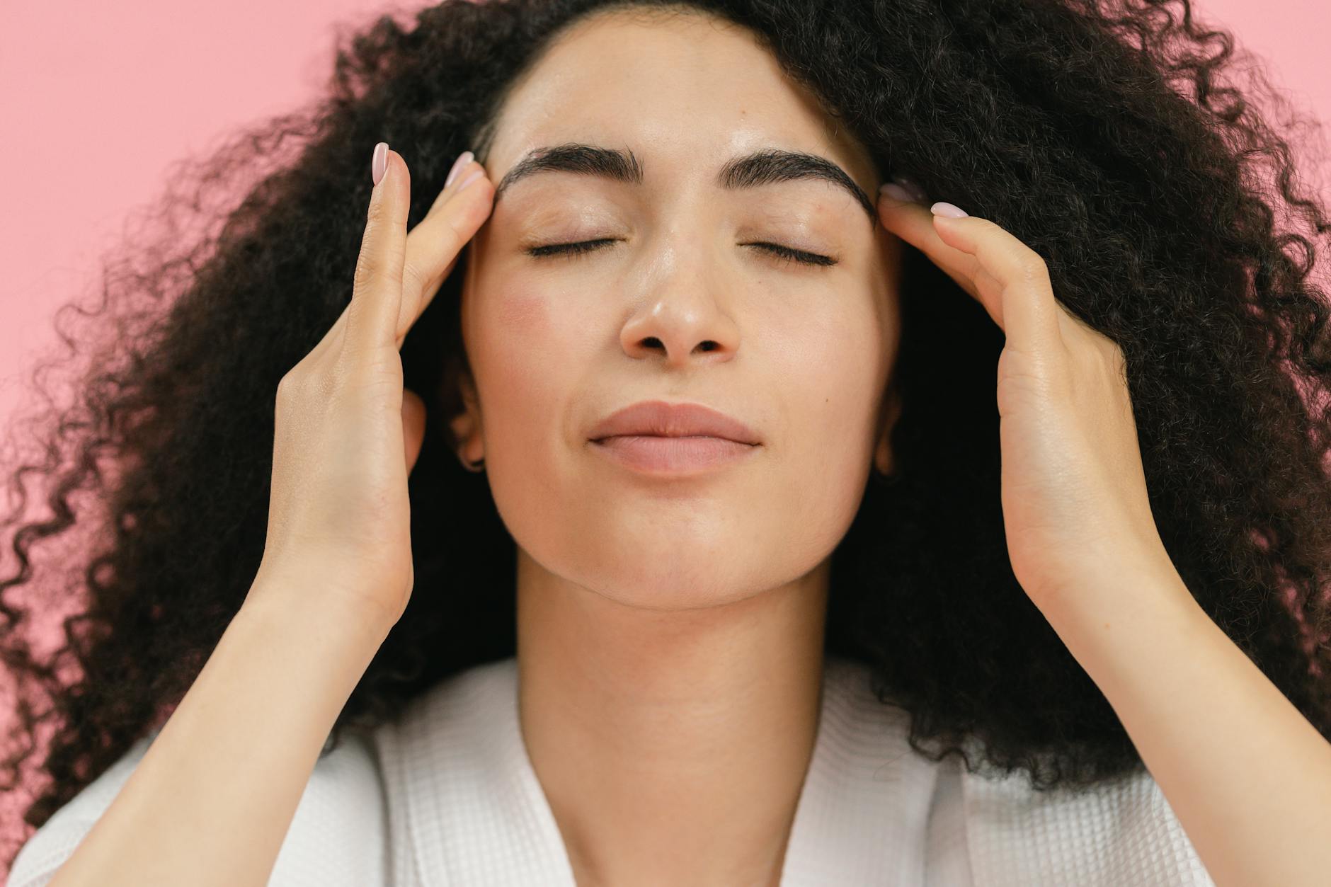 Close-up of a woman enjoying a relaxing facial massage against a pink background.