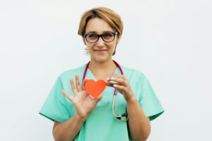 Female doctor in scrubs holding a paper heart symbol, smiling with a stethoscope around her neck.